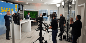 The same television studio, but this time several people are visible in the image, including camera operators and presenters. The blue wall with the text 