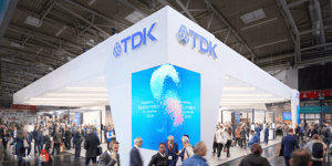 Angled view of the TDK booth featuring a large digital screen displaying colorful graphics and text, surrounded by attendees.