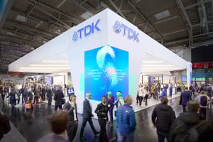 Schräger Blick auf den TDK Messestand mit großem digitalen Bildschirm, der bunte Grafiken und Text zeigt, umgeben von Messebesuchern.