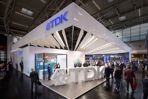 Perspektive des modernen TDK Messestandes mit auffälliger Beleuchtung und Besuchern, die sich mit den Exponaten beschäftigen.