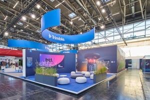 Open-plan Trimble exhibition stand with a curved overhead banner, lounge area, digital presentation displays and integrated greenery.