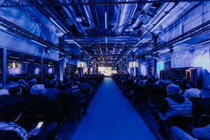 ZEISS‑Roadshow‑Präsentation in einer Industriehalle mit atmosphärischer blauer Beleuchtung und einem großen Publikum, das zur Hauptbühne blickt.