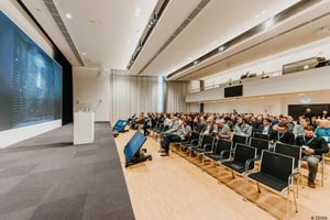 Keynote‑Session der ZEISS‑Roadshow mit einem Vortragenden auf der Bühne vor einer großen digitalen Präsentationsfläche und einem sitzenden Publikum in einem hellen Konferenzraum.
