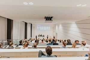 Eine ZEISS‑Präsentation in einem modernen Vortragssaal, bei der Teilnehmende den Referenten auf der Bühne folgen und projizierte Inhalte ansehen.