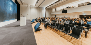 Keynote‑Session der ZEISS‑Roadshow mit einem Vortragenden auf der Bühne vor einer großen digitalen Präsentationsfläche und einem sitzenden Publikum in einem hellen Konferenzraum.