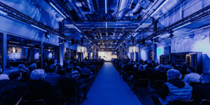ZEISS‑Roadshow‑Präsentation in einer Industriehalle mit atmosphärischer blauer Beleuchtung und einem großen Publikum, das zur Hauptbühne blickt.