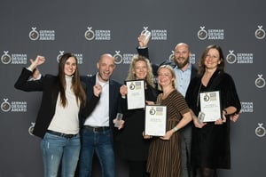 Six smiling people celebrating posing in front of a camera and holding several awards.