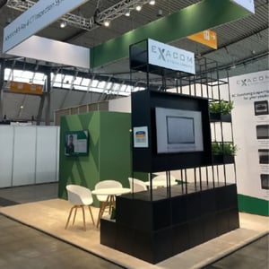 EXACOM exhibition stand with green displays, chairs and cupboards.