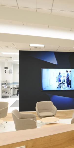 Heller Wartebereich in einem medizinischen Showroom mit bequemen Stühlen um kleine Tische, einem großen Bildschirm, auf dem medizinisches Fachpersonal mit einem Patienten im Rollstuhl zu sehen ist, und im Hintergrund sichtbaren Untersuchungsräumen.