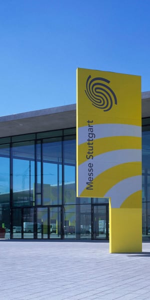 Außenansicht des Landesmessegebäudes in Stuttgart mit einem großen gelben Banner, auf dem der Schriftzug „Messe Stuttgart“ und ein kreisförmiges Logo vor strahlend blauem Himmel zu sehen sind.