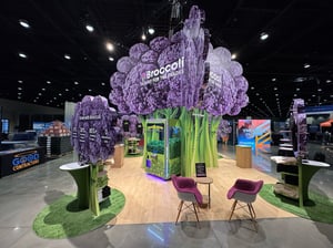 Broccoli AI trade show booth designed as a large stylized purple and green broccoli structure. A central square display area features screens and equipment. Two purple chairs sit on a green carpet section. Additional expo booths are visible in the background.