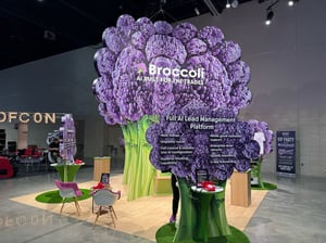 Close-up of the Broccoli AI booth featuring a large purple broccoli decoration. Visible text reads: ‘Broccoli AI – A Full Lead Management Platform.’ Two purple chairs and a small table with items are placed in front of the booth.