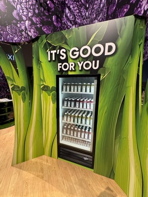 Detail view of a refrigerated display filled with bottled drinks in front of a green broccoli graphic. Above it, the text reads ‘IT’S GOOD FOR YOU.’