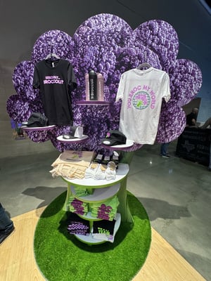 Broccoli AI merchandise display with T-shirts labeled ‘BROCCOLI AI’ and ‘BROCC MY WORLD’ hanging in front of a purple broccoli wall. A round table holds tote bags, stickers, and other branded items.