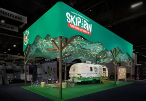 Wide view of the booth with a green overhead banner reading ‘SKIP • BIN – TRASH EDUCATION.’ Beneath it are the branded white trailer, camping chairs, a wooden table, and decorative elements including a fire pit and wooden posts.