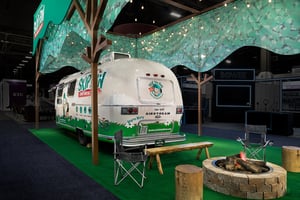 Trade show booth featuring a white retro-style trailer with green-and-white graphics and the National Recycling & Waste Association logo. Camping chairs, a wooden table, and a circular stone fire pit are arranged in front of the trailer. A green canopy resembling tree foliage is suspended above, supported by wooden posts.