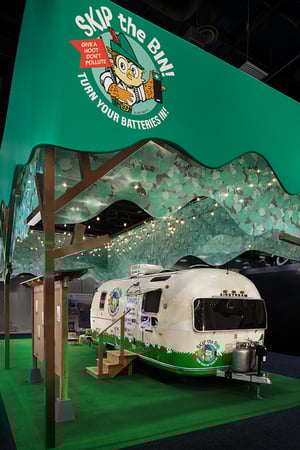 Close-up of the green, semi-transparent fabric canopy installed above the booth and held by wooden beams. Below the canopy stands the branded white trailer, illuminated by strings of lights hanging underneath the fabric.