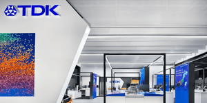 TDK booth at CES 2025 featuring a white wall with colorful abstract artwork. In the foreground, an open display area with blue screens and technology exhibits.