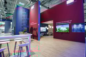 Detailansicht des Krones Messestandes mit roter Wand, Informationsbildschirmen und Stehtischen für Gespräche.